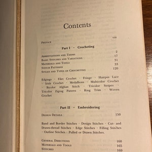 May include: Open book displaying the contents page, with sections on crocheting and embroidering. The page features a list of topics, including abbreviations, basic stitches, materials, and various crochet styles. The book has a red spine.