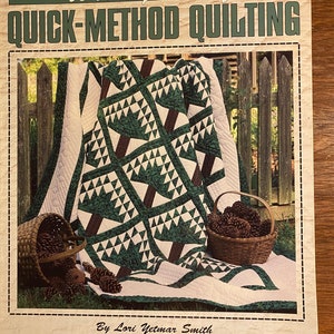 Pode incluir: Um folheto de artesanato intitulado "Ensine-se a fazer quilting de método rápido" por Lori Yetmar Smith. A capa apresenta um edredom verde e branco com um padrão geométrico, rodeado por uma cerca de madeira e pinhas em cestos.