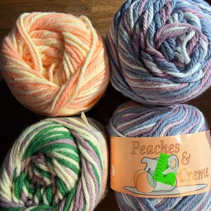 May include: Four skeins of Peaches & Creme yarn. One skein is peach and cream, one is blue, purple, and white, one is green, purple, and cream, and one is blue, purple, and white with a label featuring a peach illustration and text.