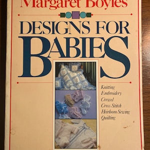 Könnte beinhalten: Ein Vintage-Buchcover mit dem Titel "Margaret Boyles' DESIGNS FOR BABIES". Das Cover hat einen cremefarbenen Hintergrund mit rotem und blauem Text. Abbildungen von Babyartikeln werden angezeigt, darunter eine Steppdecke, Babyschuhe und ein Kleidungsstück. Stichwörter: Stricken, Sticken, Crewel, Kreuzstich, Erbstücknähen, Quilten.