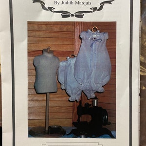 May include: A printed page featuring doll clothes patterns. The image shows a light blue doll romper and bloomers hanging on a gold hanger. A miniature sewing machine and a dress form are also visible. Text reads "The Grace Knott Doll Clothes Collection."