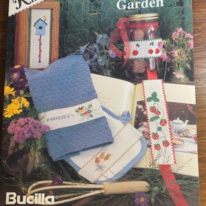 May include: A Bucilla craft book titled "A Silk Ribbon Garden" featuring cross-stitch projects. The cover displays a blue towel with "Pansies" embroidered on it, a jar with a ribbon, and a bookmark with strawberries.