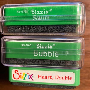 Puede incluir: Dos contenedores de plástico verde con almohadillas de espuma negra en el interior. Los contenedores están etiquetados con el nombre de la marca "Sizzix" y los nombres de las almohadillas de espuma: "Swirl", "Bubble" y "Heart, Double".