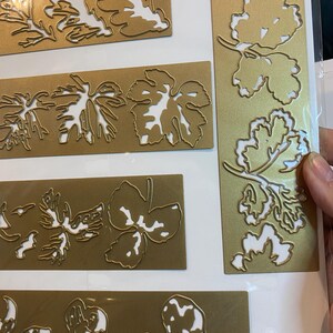 Może przedstawiać: Kolekcja złotych metalowych wykrojnik&oacute;w w kształcie liści z białą przestrzenią negatywną. Wykrojniki przedstawiają liście dębu i winorośli w r&oacute;żnych rozmiarach i kształtach. Liście ułożone są na prostokątnych i pionowych złotych tłach.