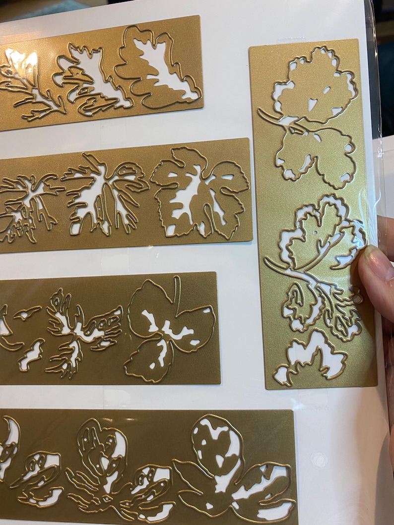 Może przedstawiać: Kolekcja złotych metalowych wykrojnik&oacute;w w kształcie liści z białą przestrzenią negatywną. Wykrojniki przedstawiają liście dębu i winorośli w r&oacute;żnych rozmiarach i kształtach. Liście ułożone są na prostokątnych i pionowych złotych tłach.