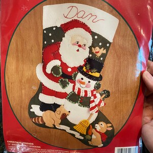 Könnte beinhalten: Ein Bucilla Christmas Friends Filzstrumpf-Set mit einem Weihnachtsmann- und Schneemann-Design. Der Strumpf ist 38 cm lang und enthält Baumwoll-Stickgarn, farblich getrennt. Das Set enthält auch Anleitungen für einfache Applikationen und Stickereien. Der Strumpf ist mit dem Namen "Dan" personalisiert.