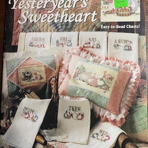 May include: A craft book titled "Sunbonnet Sue Yesteryear's Sweetheart" with cross-stitch patterns. The cover features the title and images of finished cross-stitch projects, including small embroidered towels labeled with days of the week and a pillow.