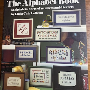 Op de afbeelding: Het boek "The Alphabet Book" van Leisure Arts, met 13 alfabetten, 6 sets cijfers en 9 randen. De omslag toont voorbeelden van kruissteekprojecten, waaronder "Tilted Block" en "Patchwork Christmas".