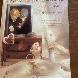 May include: Book cover with the title "Elegant Ribbon Embroidery on Felt" by Sandy Belt. The cover features a heart-shaped felt project with ribbon embroidery and buttons. The book is published by Indygo Junction, Inc.
