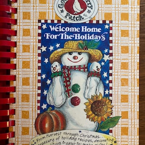 May include: A spiral-bound cookbook titled "Welcome Home For The Holidays" with a snowman illustration. The cover features a snowman wearing a straw hat and scarf, with a pumpkin and sunflower. The background has a yellow plaid pattern.