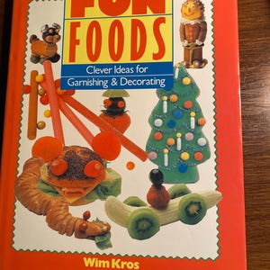 May include: A red book titled "Fun Foods: Clever Ideas for Garnishing & Decorating" by Wim Kros. The cover features colorful illustrations of food-based crafts, including a gingerbread man, a Christmas tree made of candy, a caterpillar made of bread, and a banana car.