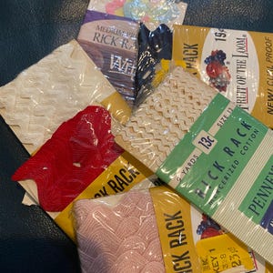 Op de afbeelding: Assortiment van vintage Rick Rack-sierband in diverse kleuren, waaronder wit, rood en roze, verpakt in transparante plastic. Sommige verpakkingen tonen tekst zoals "Fruit of the Loom" en "Mercerized Cotton".