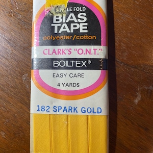 Op de afbeelding: Een verpakking met gele enkelvoudige biaisband, gemaakt van polyester en katoen. De verpakking is gelabeld met "Clark's "O.N.T." Boiltex® Easy Care" en "182 Spark Gold". Het bevat 4 yards band.
