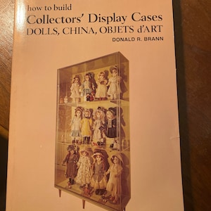 Puede incluir: Un libro titulado "Collectors' Display Cases Dolls, China, Objects d'Art" de Donald R. Brann. La portada del libro presenta una fotografía de una vitrina de vidrio llena de muñecas de porcelana.