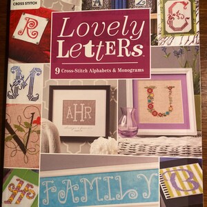 May include: A book cover for a cross-stitch pattern book titled "Lovely Letters". The cover features a variety of cross-stitch designs of letters and monograms, including the letters R, M, U, and the monogram AHR. The book cover also features the text "9 Cross-Stitch Alphabets & Monograms".