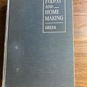 May include: A vintage book titled "Foods and Home Making" by Greer. The book has a blue cover with white text. The title is in a rectangular box. The book is on a wooden surface.