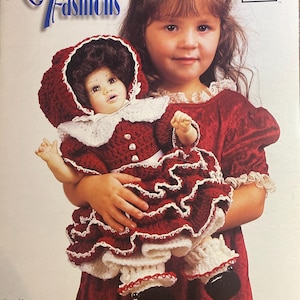 May include: A girl holding a doll dressed in a red crocheted outfit with white edging. The doll's outfit matches the cover of the "Katina's Fashions" crochet pattern book. The book cover features the text "Katina's Fashions" in blue script.