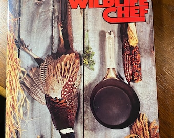 May include: The cover of a cookbook, "WILDLIFE CHEF", shows a pheasant hanging on a wooden wall. A frying pan, corn, and vegetables are also displayed. The image has a rustic, outdoorsy theme, with a focus on game cooking.