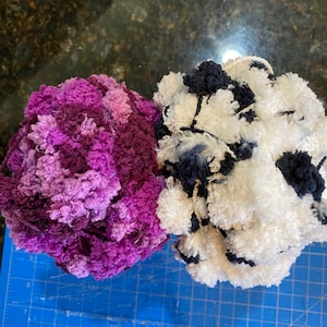 May include: Two balls of yarn, one purple and one white with black accents. The yarn is fluffy and textured.