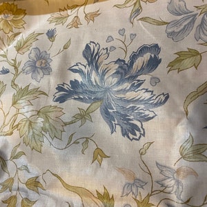 May include: Close-up of a floral fabric with a cream-colored background. The fabric features large blue and cream-colored flowers, with green and tan leaves and stems. The fabric has a slight sheen.