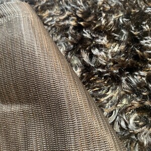 Puede incluir: Primer plano de una tela marrón texturizada con un patrón tejido, parcialmente enrollada, que revela un material rizado contrastante, marrón oscuro y gris. La imagen destaca las diferentes texturas y colores de los materiales.