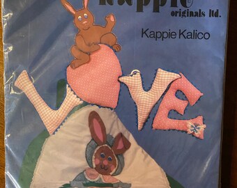 May include: A craft kit for making a wall hanging featuring the word "LOVE" in pink and white fabric with two bunnies. The kit includes patterns, directions, trim, and assorted fabrics in colors and quantities required for completion. The finished size is 15 inches by 25 inches.  The kit is from Kappie Originals Ltd.