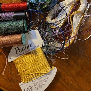 以下が含まれることがあります： 金、赤、白、青など、さまざまな刺繍糸の糸巻きをクローズアップで撮影したものです。糸巻きには「Patina Needlepoint 13 to 18 Count & Cross Stitch」とラベルが貼られています。