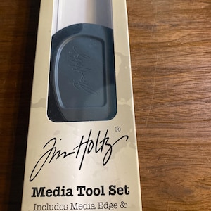 May include: A tan package for a Jim Holtz Media Tool Set. The package features black text, including "Media Tool Set", and a clear window revealing the media edge and scraper. The tools are designed for the Tim Holtz Glass Media Mat.