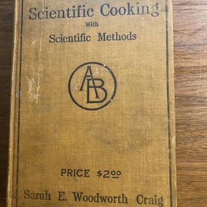 May include: A vintage cookbook titled "Scientific Cooking with Scientific Methods" by Sarah E. Woodworth Craig. The cover is a faded tan color with black text and a circular logo. The price is listed as $2.00.