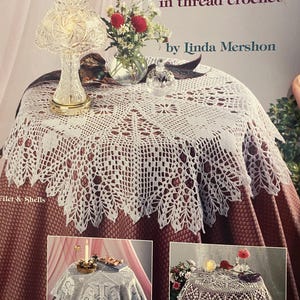 以下が含まれることがあります： リンダ・マーション著「ロマンチックテーブルトッパーズinスレッドかぎ針編み」の表紙。ブルゴーニュ色のテーブルクロスの上に白いかぎ針編みのテーブルトッパー、クリスタルランプと花が描かれています。
