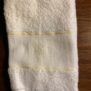 Puede incluir: Toalla blanca doblada con una superficie de rizo texturizada. La toalla presenta una banda tejida con una raya amarillo claro y flecos en el borde inferior. Adecuada para uso en ba&ntilde;o o spa.