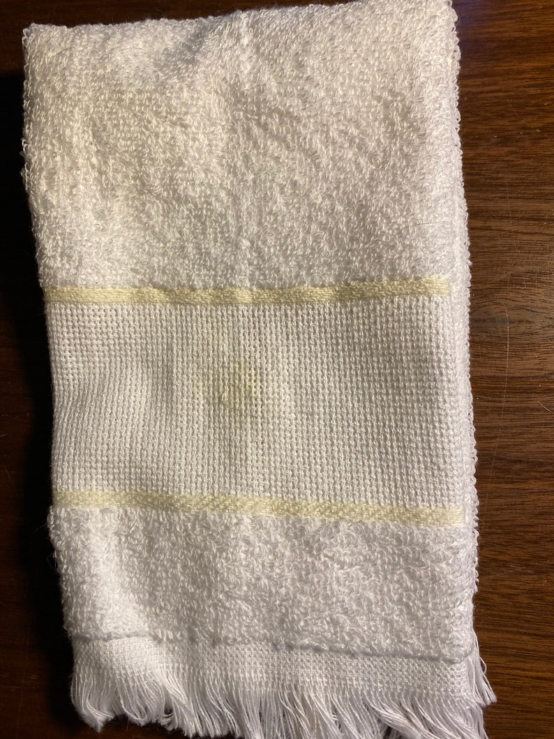 Puede incluir: Toalla blanca doblada con una superficie de rizo texturizada. La toalla presenta una banda tejida con una raya amarillo claro y flecos en el borde inferior. Adecuada para uso en ba&ntilde;o o spa.