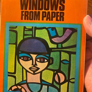 May include: Book cover with the title "Stained Glass Windows From Paper" in black text on an orange background. The cover features a colorful stained glass illustration of a person with a blue hat and a bird.