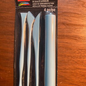 May include: A set of four blue plastic clay sculpting tools in a clear plastic package. The package has the text "craft smart" and "PREMIUM CLAY TOOL SET" on it. The tools include a round tool, a pointed tool, a flat tool, and a curved tool.