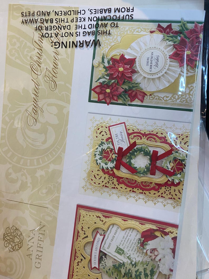 Może przedstawiać: Opakowanie warstwowych wykrojnik&oacute;w kwiat&oacute;w bożonarodzeniowych. Wykrojniki przedstawiają czerwone poinsecje, wieńce i dekoracyjne ramki. Opakowanie zawiera tekst "Sending Christmas Wishes" i "Season's Greetings".