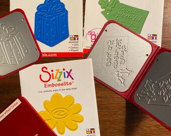 May include: Three Sizzix Embosslits™ die cut templates for crafting. The first die is a blue gift box with a bow. The second die is a green tag with the text "Don't open 'til Dec. 25". The third die is a yellow flower with a swirl in the center.