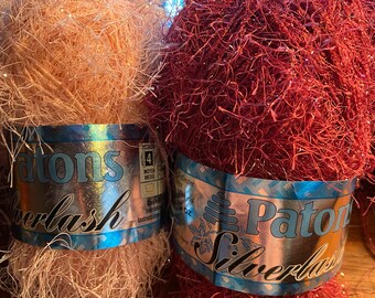 Può includere: Due gomitoli di filato Patons SilverLash, uno di colore rosa pesca e l'altro rosso bordeaux scuro. Entrambi i gomitoli hanno una consistenza soffice e scintillante.