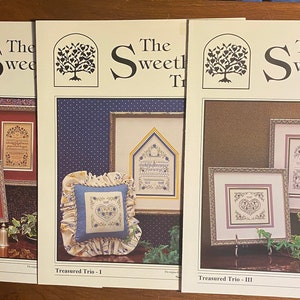May include: Three framed cross-stitch patterns titled "Treasured Trio" with the text "The Sweetheart Tree." Each frame features a different design, including hearts and floral motifs. The frames are in red, gold, and silver.