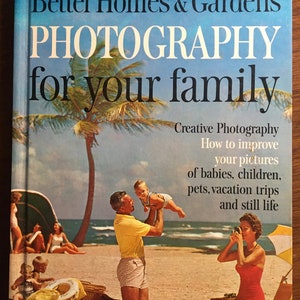 Puede incluir: Un libro titulado "Better Homes & Gardens Fotografía para tu familia" con una imagen de portada de una familia en una playa. La familia está compuesta por un hombre que sostiene a un niño pequeño en el aire, una mujer que toma una foto y dos niños sentados en la arena. La portada del libro también incluye el texto "Fotografía creativa, Cómo mejorar tus fotos de bebés, niños, mascotas, viajes de vacaciones y naturalezas muertas."