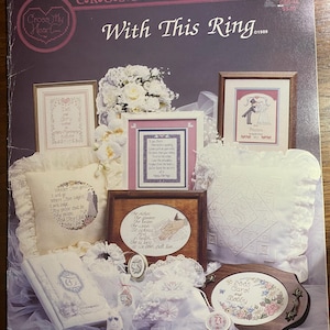 Może przedstawiać: Vintage książka z wzorami haftu krzyżykowego zatytułowana "With This Ring" autorstwa Cross My Heart, Inc. Okładka zawiera oprawione próbki haftu krzyżykowego, poduszki i elementy dekoracyjne o tematyce ślubnej. Wzory obejmują motywy kwiatowe i romantyczne.