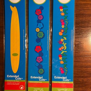 May include: Three packages of Ellison Design Extended Cuts die-cutting templates. The first package features a surfboard design, the second package features buttons, and the third package features flowers. Each package is blue with white text and a small image of the design. The text on the packages reads "Extended Cuts" and includes the name of the design and the designer. The packages are compatible with Tag-a-long, Big Shot, and other die-cutting systems.