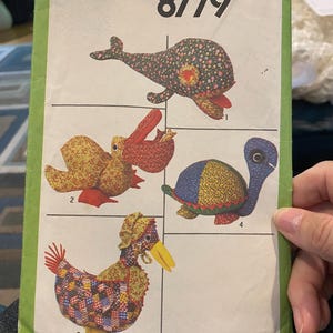 Op de afbeelding: Simplicity naaipatroon nummer 8779 voor het maken van vier knuffels: een walvis, een eend, een schildpad en een kip. Het patroon is gedrukt op een witte achtergrond met groene randen.