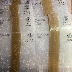 Puede incluir: Una colección de carpetas de relieve Cricut Cuttlebug de Anna Griffin. Las carpetas son de plástico transparente con varios diseños en relieve, incluyendo patrones florales, formas geométricas y letras.