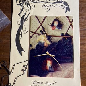 May include: A craft pattern booklet titled Country Heartstrings featuring a photograph of three small fabric angel ornaments with wire wings. The text reads Littlest Angel and 2 1/2" Tall - Wire wings included. A single wire angel wing frame is visible.