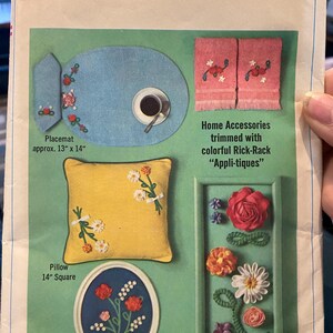 May include: A Simplicity sewing pattern for home accessories, featuring a blue placemat, a yellow pillow, a framed floral motif, and a framed arrangement of colourful rick-rack flowers. The pattern is titled "Simplicity 6805" and is priced at 50 pence.