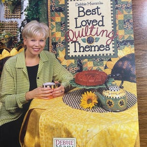 Puede incluir: Portada del libro "Debbie Mumm's Best Loved Quilting Themes" con una mujer sonriendo, sosteniendo una taza. La portada tiene un diseño temático de edredones con girasoles y una tetera decorativa. La mesa está cubierta con un mantel amarillo.