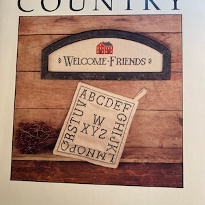 Op de afbeelding: Een houten bord met een rode huisillustratie en de tekst "Welcome Friends" in zwarte letters. Een witte stof met het in zwart geborduurde alfabet ligt op een houten oppervlak.