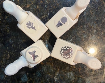 May include: Four white paper punches with various designs: a leaf, an ice cream cone, a butterfly, and a floral pattern. Each punch has a handle for easy use. The punches are arranged in a cross formation.