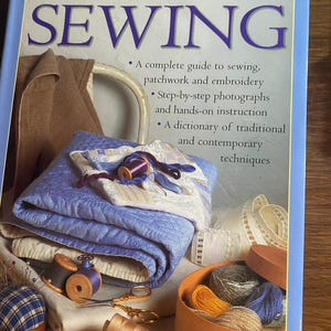 May include: A hardcover book titled The Practical Encyclopedia of Sewing by Dorothy Wood. The cover features a light blue border, a quilted lavender fabric, wooden spools of thread, metal scissors, a pincushion, and a round box of yarn.
