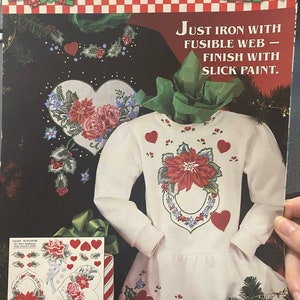 Puede incluir: Una camisa blanca con un diseño de poinsettia roja y corona de acebo. La camisa está decorada con corazones rojos y hojas de acebo verdes. La imagen incluye instrucciones para aplicar el diseño a la camisa usando tela fusible. El texto en la imagen dice "Daisy Kingdom No-Sew Applique #6390 Holly Hearts Just Iron With Fusible Web - Finish With Slick Paint."